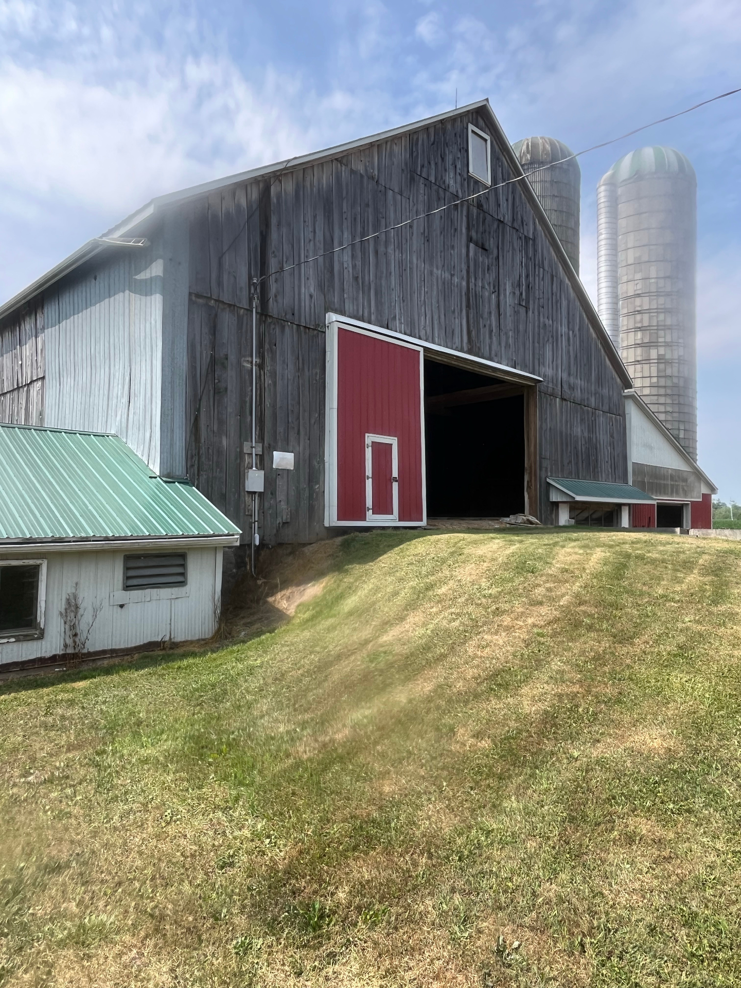 Weathered grey board-and-batten bank barn with red sliding door and twin silos before re-cladding