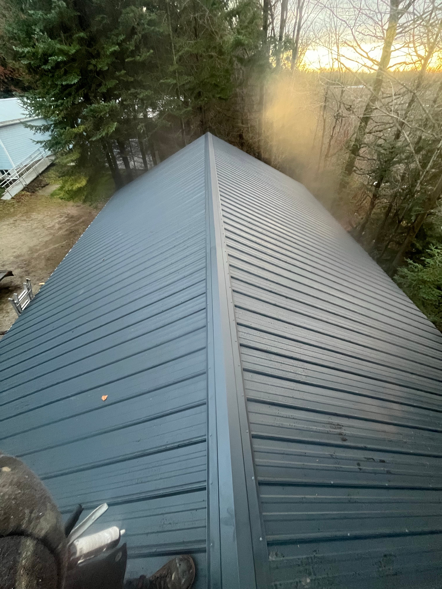 Freshly installed dark metal roof with a clean center ridge line beside a wooded property at sunset
